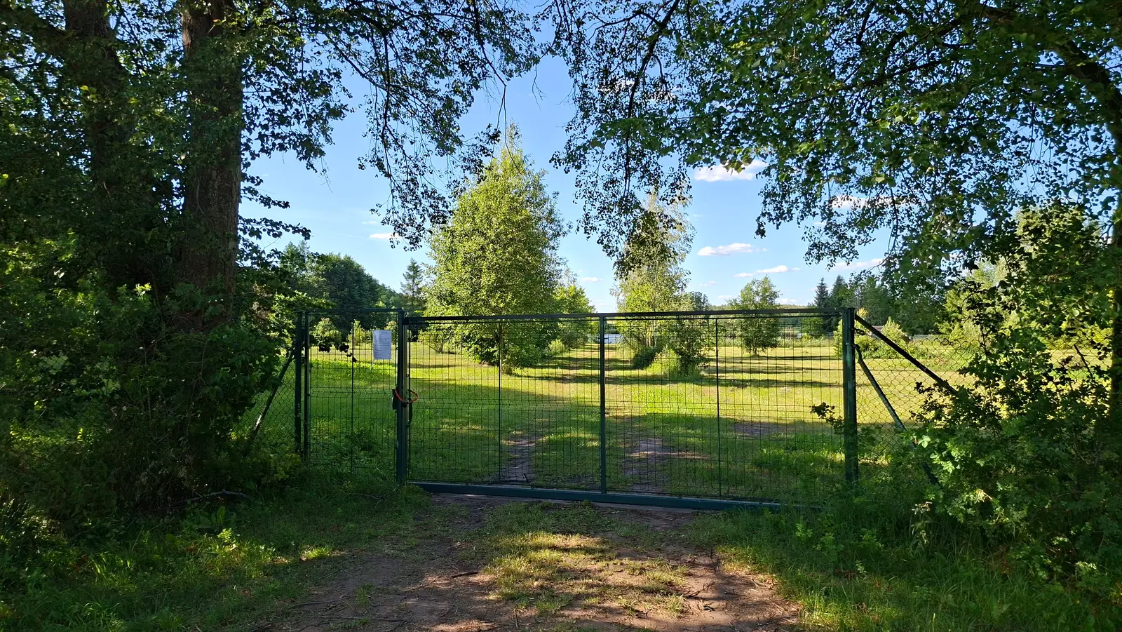 Krajobraz w formacie poziomym przedstawia metalową, zieloną bramę wjazdową na ogrodzony teren zielony, otoczony gęstymi drzewami i krzewami. Centralny punkt zdjęcia stanowi prostokątna brama wykonana z siatki, przez którą widać nasłonecznioną polanę z młodymi drzewami pod błękitnym, bezchmurnym niebem. Na lewym skrzydle bramy widoczna jest biała kartka z wydrukowanym tekstem, której treść jest nieczytelna z tej odległości. Nad sceną rozciągają się gałęzie drzew, które tworzą naturalną ramę i rzucają cień na piaszczystą ścieżkę prowadzącą do wejścia. Kontrast między cienistym pierwszym planem a jaskrawo oświetlonym tłem podkreśla głębię obrazu i sielski, letni klimat miejsca.